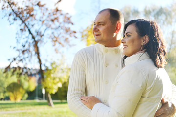 Fototapeta premium Husband and wife in white warm clothes pose for the camera in an autumn park on a sunny day. couple looking away