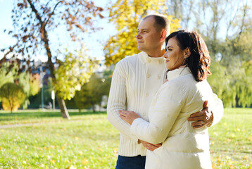 Husband and wife in white warm clothes pose for the camera in an autumn park on a sunny day. couple looking away