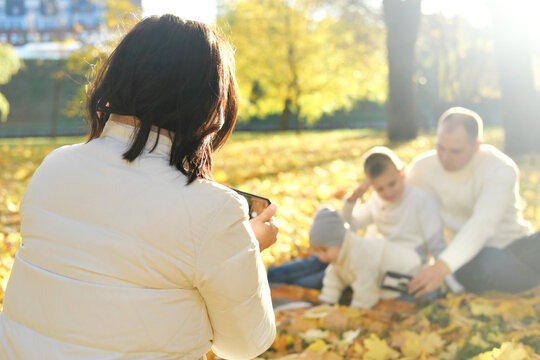A happy family of four spend time in the autumn park. Mother taking pictures of her family or recording video on mobile phone