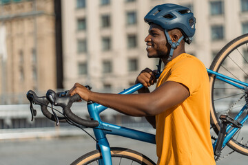 Portrait of handsome smiling African American man wearing helmet holding his bicycle walking on urban street looking away. Sport, hobby, active lifestyle concept 