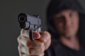 robber with gun aiming into camera. Man in hood threatens with firearm. Weapon in person's hands....