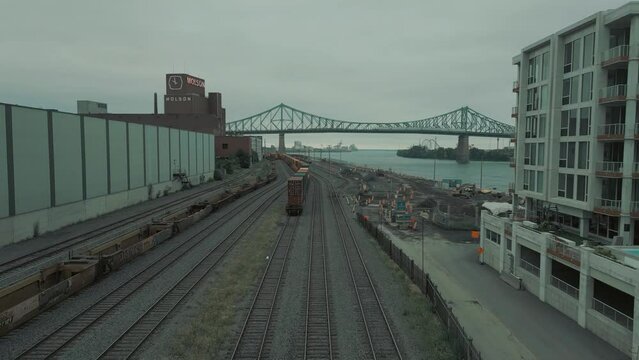 Forward pan towards Jaque Cartier Bridge in Montreal then face down towards train on train tracks before the bridge with a factory and bright red signage 