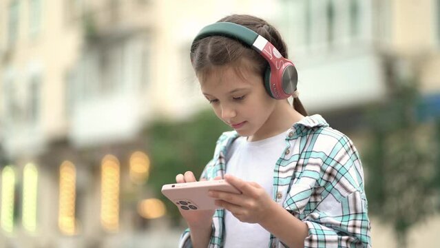 Child girl listens to music in headphones through an online smartphone application outdoors.