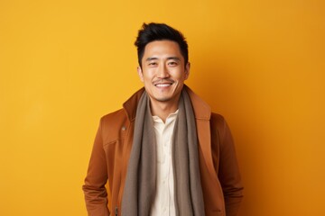 Fototapeta premium Smiling young asian man wearing coat and scarf over yellow background