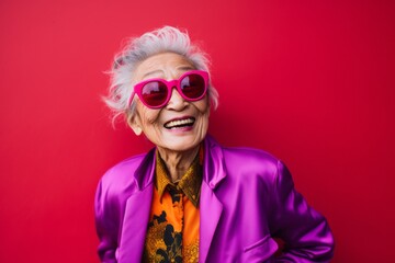 stylish senior woman in pink jacket and sunglasses smiling at camera isolated on red