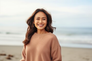 happy asian woman in beige sweater looking at camera on beach