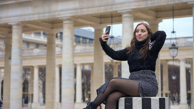 Delighted Latina woman traveler smiling and taking self portrait on smartphone while sitting on white and black pillar near Palais Royal in Paris, France
