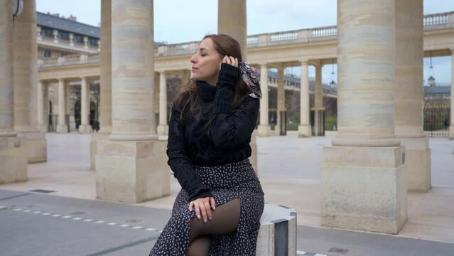 Delighted Latina woman traveler smiling while sitting on white and black pillar near Palais Royal in Paris, France