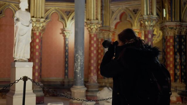 Side view of young Hispanic woman in warm jacket taking photo with professional camera of majestic interior of the Gothic chapel Sainte Chapelle in city of Paris, France