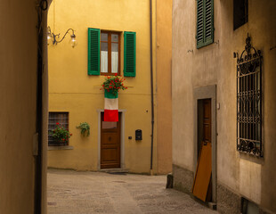 Walkway between buildings in small Italian village located in Emilia-Romania region in Northern Italy.  Brightly painted buildings with Italian flag and window shutters and street lights - obrazy, fototapety, plakaty