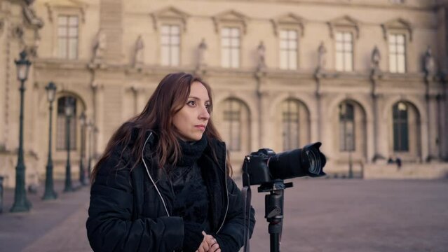 Positive Latina woman photographer in warm clothes taking photos on professional photo camera on tripod or stationary holder while standing near Louvre museum in Paris, France