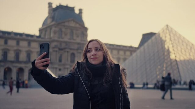 Positive young Latina woman blogger broadcasting online on social media and shooting video on smartphone against historical Louvre museum in Paris, France during sunset