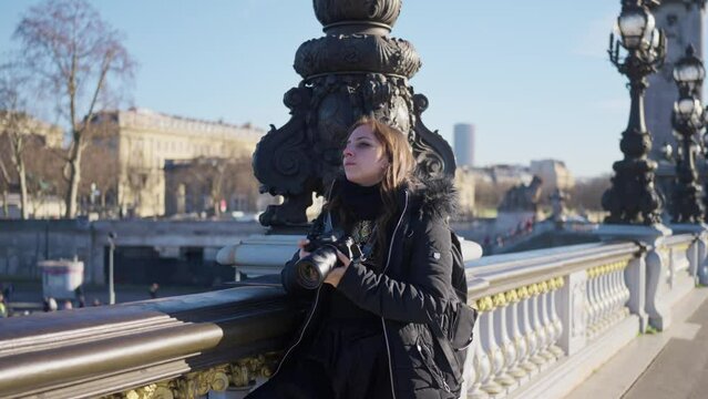 Thoughtful and pensive young Latin woman traveler in warm outfit standing on Pont Alexandre bridge with professional photo camera and looking away during sightseeing trip in Paris, France