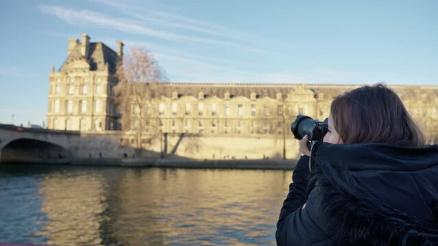 Smiling young Latina woman in warm clothes taking photos while having boat trip on river Seine with aged bridge in Paris, France