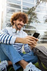 Happy young male college student sitting on grass in campus using mobile phone. Vertical image.