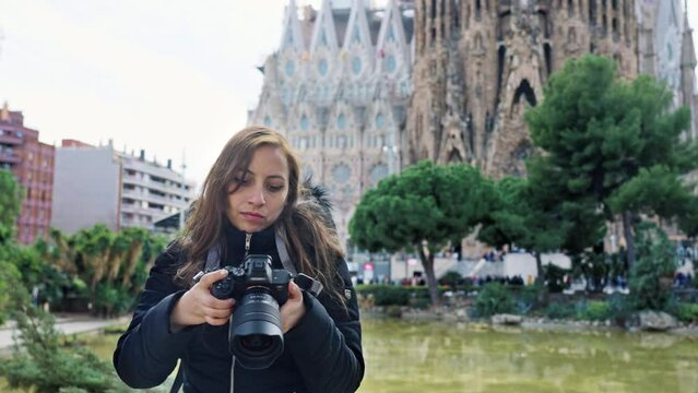 Calm young Latin woman tourist in warm outfit with photo camera looking at camera while standing against amazing church Sagrada Familia by Gaudi during sightseeing trip through Barcelona, Spain