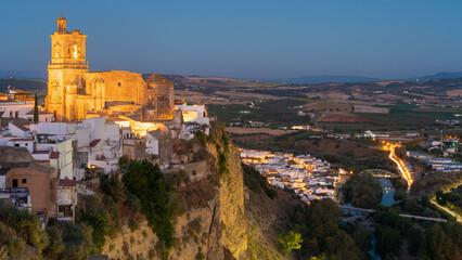 Village of Arcos de la Frontera, Spain, during the blue hour.  Iglesia de san pedro is bathed in golden light perched on a cliff with a cobalt blue sky background.