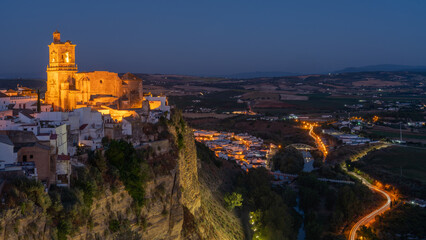 Fototapeta na wymiar Village of Arcos de la Frontera, Spain, during the blue hour. Iglesia de san pedro is bathed in golden light perched on a cliff with a cobalt blue sky background.