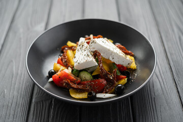 Healthy greek salad of fresh vegetables with tomatoes, bell pepper, cucumbers, olives, feta cheese and sun dried tomatoes in plate on black wooden background. Healthy food, vegetarian dieting