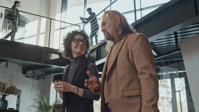 Medium shot of middle-aged Caucasian businessman and female colleague in glasses and smart casual clothes walking together through office building lobby, man talking and woman smiling and listening
