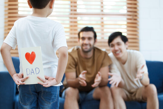 Asian lovely little boy happy to give his dad a Father's Day greeting card and present while living together in living room, diverse gender and lifestyle concept.