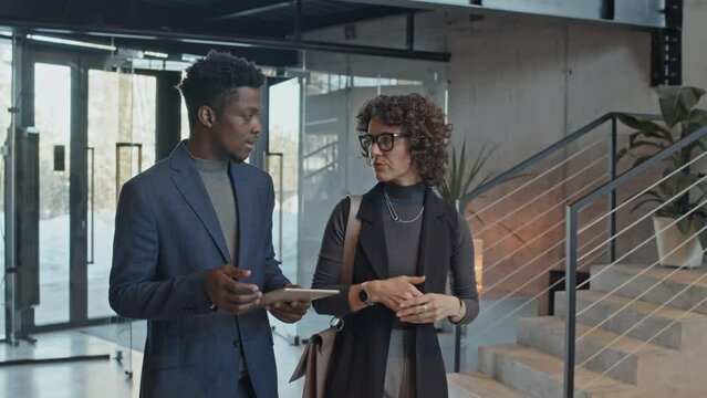 Medium follow shot of young African American junior associate with tablet computer talking to Caucasian female colleague on way to work, sharing difficulty and asking for advice and opinion