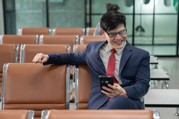 Naklejka na ściany i meble Businessman sitting on chair meeting remotely via mobile phone. Man smiling happily while playing social media. Businessman waiting to board the plane inside the airport.Front view of an Asian man.