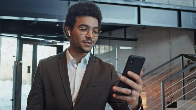 Medium close-up shot of young Middle Eastern man in glasses and casual suit, wearing wireless earphones holding up smartphone and speaking on video call on way to office