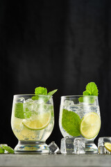 Two glasses of mojito cocktail with mint leaves and lime pieces and with ice on a dark background. Vertical shot. Indoors from a low angle.