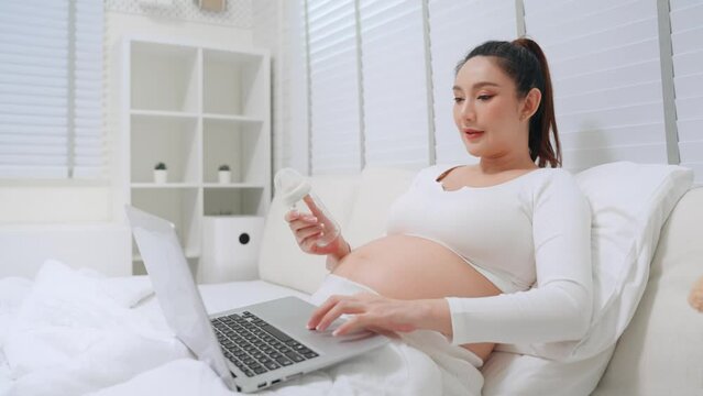 Happy pregnant asian woman use computer laptop shopping online and selection baby bottle on bed in bedroom. Asian mother preparing things baby items before prenatal while maternity leave stay at home.