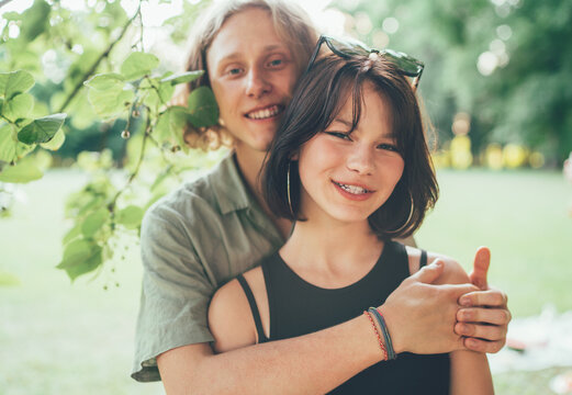 Young teenage boy hugging his girlfriend cheerfully smiling at the camera during an outdoor walk in a summer city park. First love, modern teenagers relations concept image.