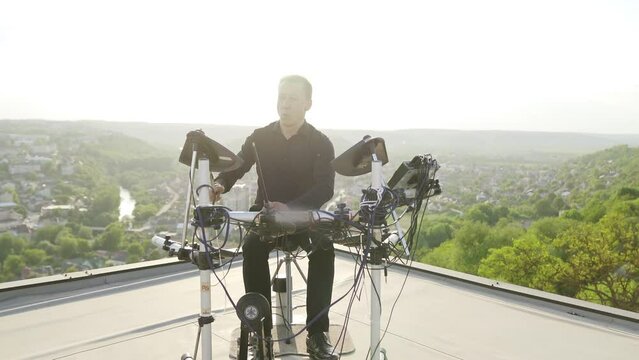 A man in black clothes plays electronic drums on a rooftop with a cityscape view