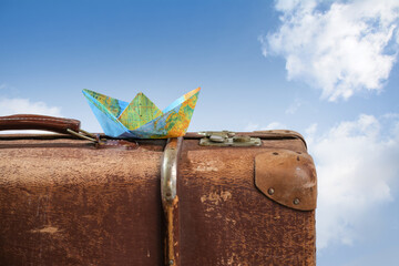 Paper boat folded from a map on an old vintage suitcase against a blue sky with white clouds, wanderlust, cruise and travel concept, copy space, selected focus