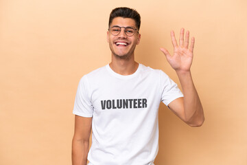 Young volunteer caucasian man isolated on beige background saluting with hand with happy expression