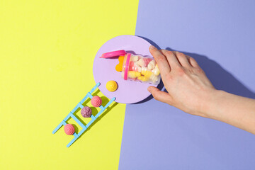 Jelly candies in jar, stepladder and female hand on yellow-purple background, top view