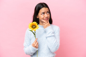 Young Colombian woman holding sunflower isolated on pink background having doubts