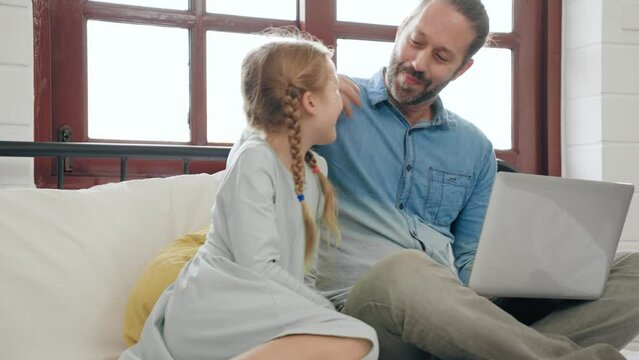 Young father working at home on computer laptop on bed while cute little daughter walking to hug with love. Relationship family concept
