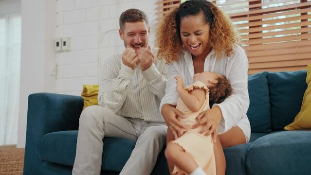 Happy parents play peekaboo and teasing with little daughter in living room at home. Father and mother using spare time with baby together at warmth house. Family relationship concept.