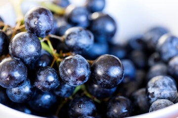 A portrait full of multiple fresh blue or black grapes in a white bowl. The fruit bunch is healthy and a great small snack to eat during a break or during a party with friends or family.