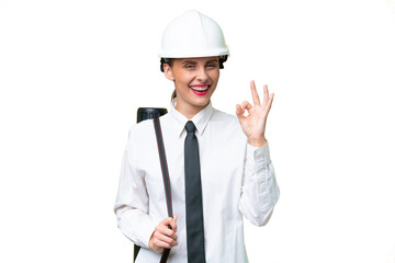 Young architect woman with helmet and holding blueprints over isolated background showing ok sign with fingers