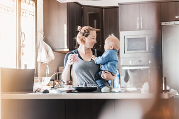 Happy mother and little infant baby boy together making pancakes for breakfast in domestic kitchen. Family, lifestyle, domestic life, food, healthy eating and people concept