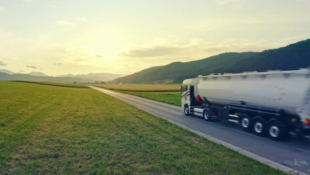 White tanker truck traveling on sunlit highway road aerial shot. Lorry transporting freight while riding through green field with mountains in background