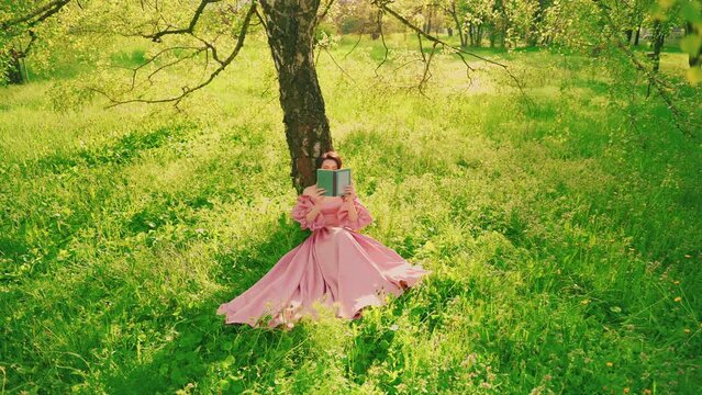 Retro woman sitting under tree reading paper book. Summer garden park green grass spring nature magic sun light, Lady holding novel art hides face behind book. long pink dress old style. girl model 4k
