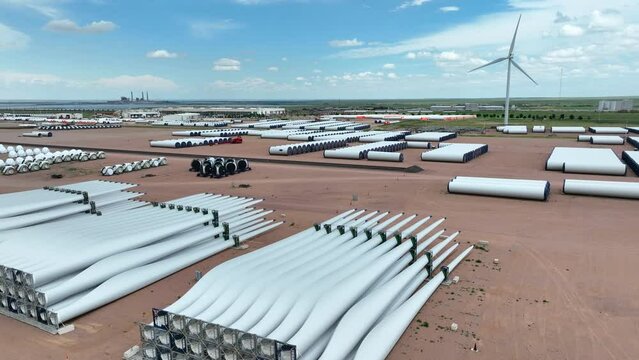 Wind turbine blades and windmill parts at large manufacturing plant. Aerial rising shot with wind turbine spinning in background.