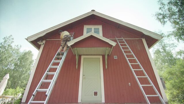 Handyman on ladder removes old red falu paint from Swedish building exterior