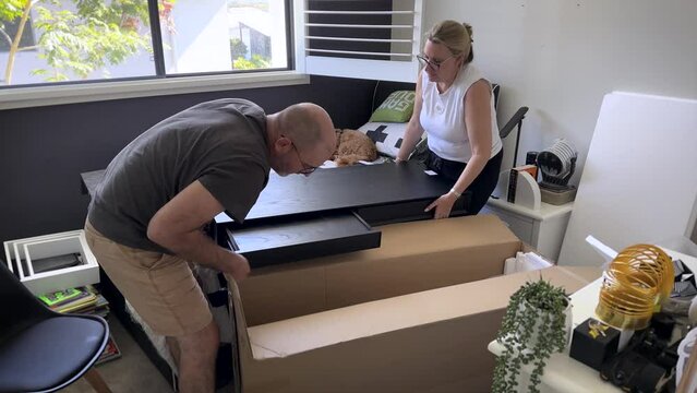 A mature couple lifts a flat-pack heavy desk to find a mark or scratch.