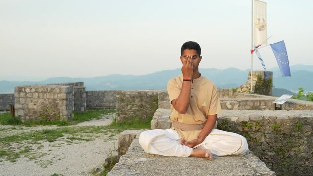 Slow-motion of young Indian male doing nadhi shuddhi meditation at sunrise on stone wall