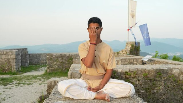 Slow-motion of Asian Indian man doing nadhi shuddhi meditation at sunrise on stone wall on top of the hill