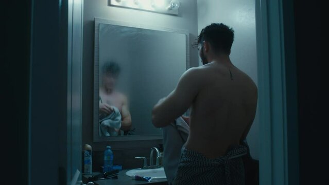 Man walks out towards mirror, towel wrapped around, wipes steam off and views his reflection in the mirror.