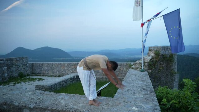 wide-shot of adult Indian man starching before doing hatha yoga barefooted in traditional yogi clothes at the top of the hill on castle wall at surise at dawn